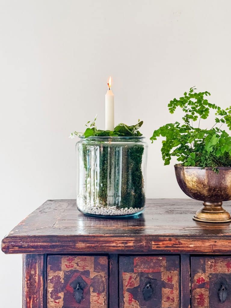 A lit candle sits on a glass container filled with moss and small plants, atop a wooden table. Beside it is a brass bowl holding a lush green plant. The background is a plain, light-colored wall.