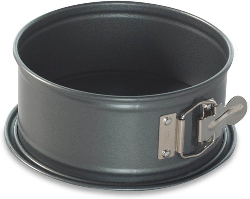 A metal springform cake pan with a smooth, dark gray finish is perfect for crafting the best spring cakes. It features a side latch for easy removal of baked goods and sits elegantly against a white background.