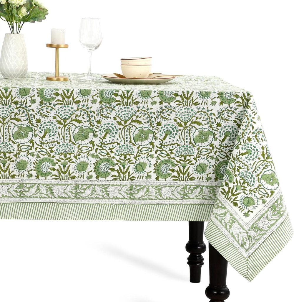 A rectangular table with a green and white floral patterned tablecloth, perfect for spring kitchen decor. On top are a vase with flowers, a candlestick, two wine glasses, and stacked dishes. The table has dark legs.