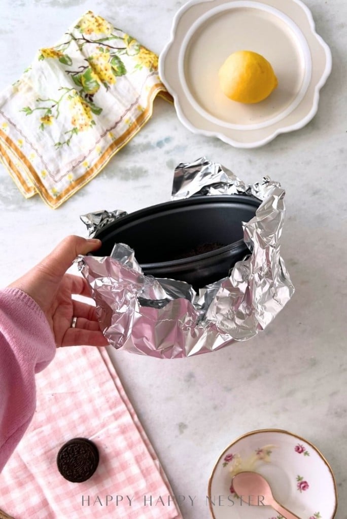 A person is holding a baking pan wrapped in aluminum foil. Nearby are a lemon on a plate, a floral towel, and hints for an Oreo Strawberry Cheesecake Recipe—a small container with dark contents and a teacup with a spoon. The setting has a light, airy kitchen feel.