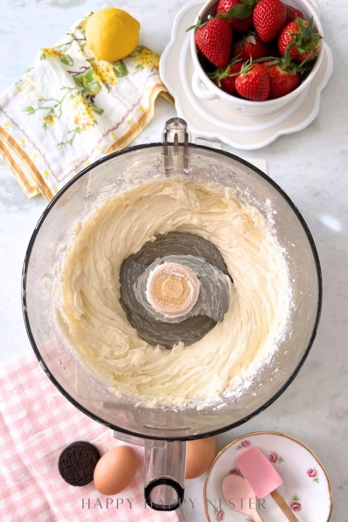 A food processor bowl with creamy batter sits on a countertop, hinting at an Oreo Strawberry Cheesecake recipe. Nearby are a lemon, a bowl of strawberries, a floral towel, a pink checkered cloth, a pink spatula, two eggs, and tempting chocolate cookies.