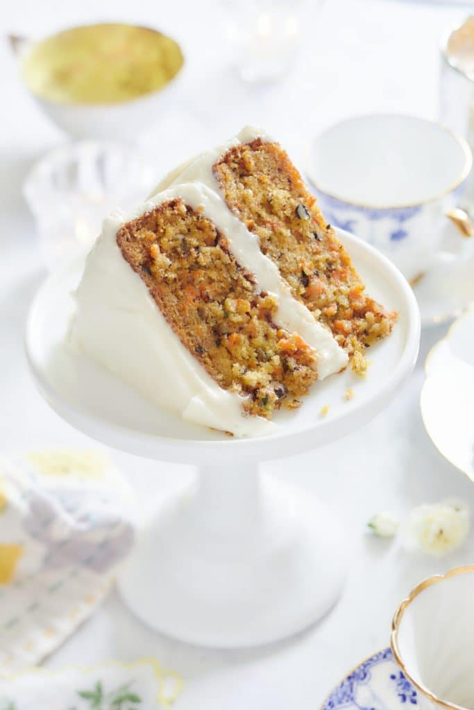 A slice of carrot cake with cream cheese frosting graces a white pedestal cake stand, epitomizing one of the best spring cakes. Delicate china cups and saucers with a blue and gold pattern adorn the light background, creating an elegant scene.