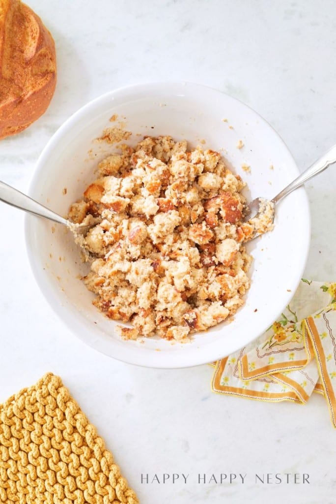 A bowl of homemade sourdough stuffing mixed with herbs rests on a marble countertop. Two serving utensils are nestled in the bowl. Nearby, a loaf of bread, a patterned cloth, and a yellow potholder complete the cozy scene. The corner text reads "Happy Happy Nester.