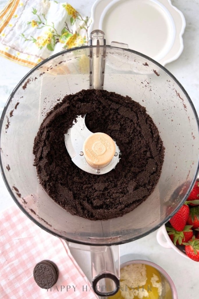 A food processor with crushed Oreo cookies inside. Surrounding it are a plate, a checkered cloth, strawberries in a bowl, and a bowl with melted butter—perfect setup for an Oreo strawberry cheesecake recipe.