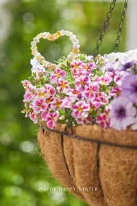 A hanging basket filled with vibrant pink, white, and purple flowers features a charming beaded heart and beaded garden stakes DIY decorations nestled among the blooms. The background is blurred greenery.