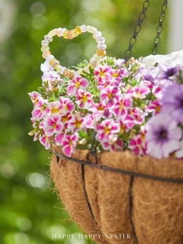 A hanging basket filled with vibrant pink, white, and purple flowers features a charming beaded heart and beaded garden stakes DIY decorations nestled among the blooms. The background is blurred greenery.
