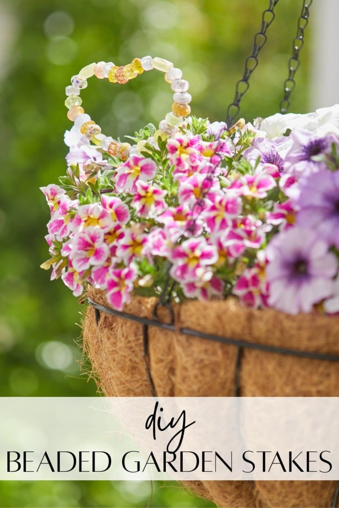 A hanging basket overflows with pink and white flowers, featuring a heart-shaped beaded garden stakes DIY accent crafted from colorful beads. Text at the bottom reads "DIY Beaded Garden Stakes.