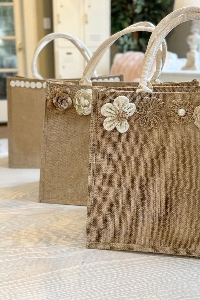 Three burlap tote bags with cream-colored handles, each decorated with fabric flowers and pearls along the top edge, are arranged on a light wooden surface in a softly lit room—perfect inspiration for your next DIY garden crafts project.