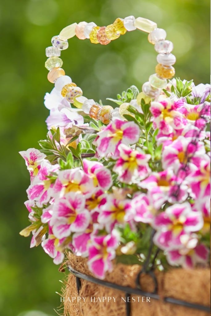 A beaded heart-shaped ornament, reminiscent of beaded garden stakes diy projects, nestles among vibrant pink and white flowers in a hanging basket with a blurred green background.