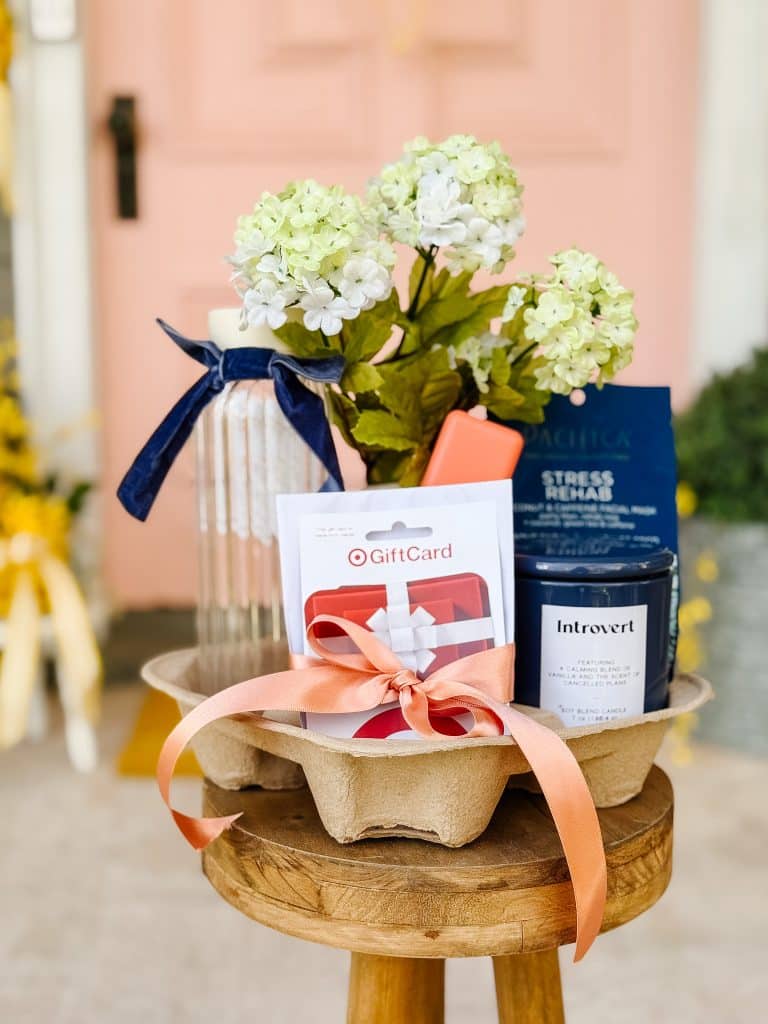 A gift basket on a wooden stool includes white flowers, a water bottle with a blue ribbon, a Target gift card, DIY Garden Stakes, a candle labeled "Introvert," and wellness products, all tied with an orange ribbon bow.