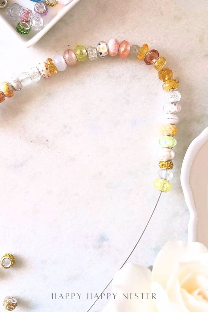 A partially strung necklace with colorful glass beads rests on a light marble surface, hinting at beaded garden stakes DIY inspiration. A white flower and a dish with extra beads sit beside it. The words "HAPPY HAPPY NESTER" appear at the bottom.