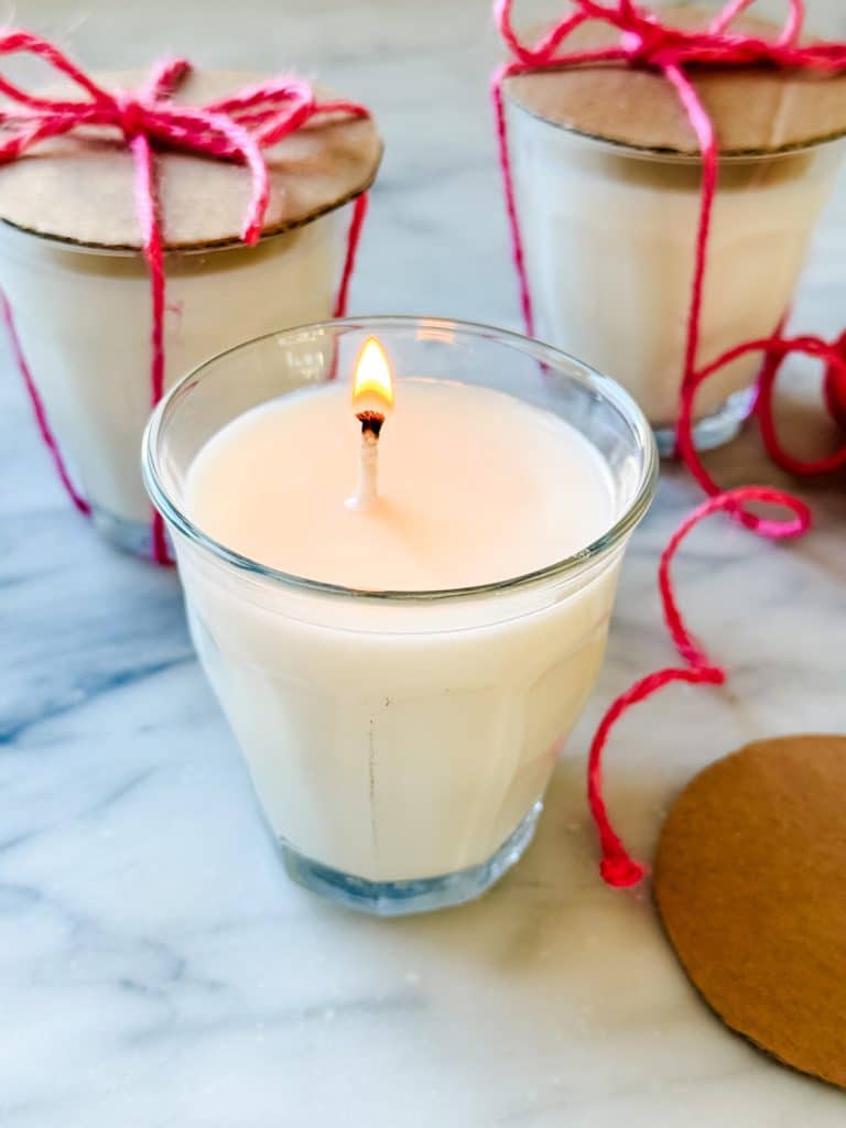 A lit white candle in a glass jar sits on a marble surface, with two more similar candles in the background. The background candles have cardboard lids tied with bright pink string, perfect for adding DIY garden decor charm to your space.