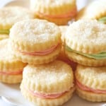 A plate of round, sugar-topped sandwich cookies filled with pastel pink and green cream, neatly stacked and displayed on a white dish, showcases a delightful cream wafer cookie recipe.