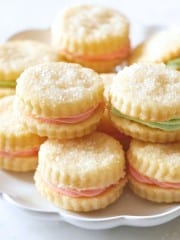 A plate of round, sugar-topped sandwich cookies filled with pastel pink and green cream, neatly stacked and displayed on a white dish, showcases a delightful cream wafer cookie recipe.