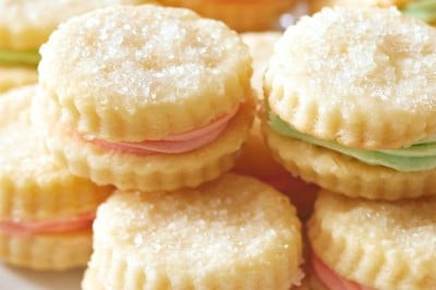 A plate of round, sugar-topped sandwich cookies filled with pastel pink and green cream, neatly stacked and displayed on a white dish, showcases a delightful cream wafer cookie recipe.