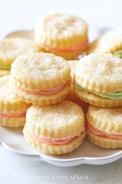 A plate of round, sugar-topped sandwich cookies filled with pastel pink and green cream, neatly stacked and displayed on a white dish, showcases a delightful cream wafer cookie recipe.
