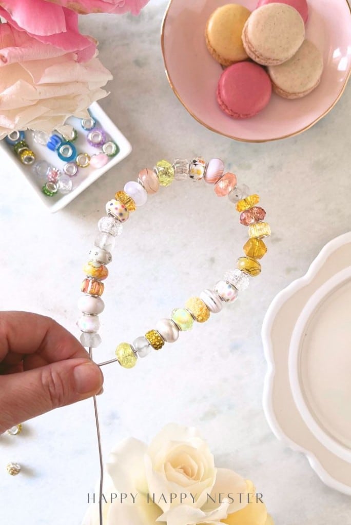 A hand holds a wire with colorful glass beads shaped into a heart. On the table are pastel macarons, flowers, small decorative beads, and plates, all arranged on a light surface.
