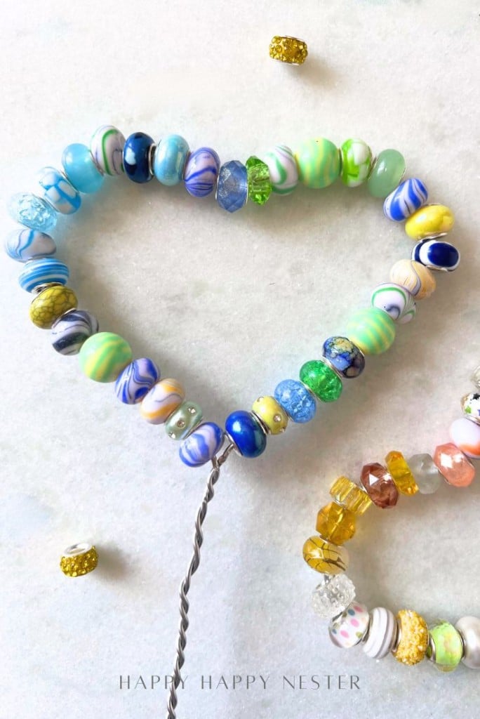 A heart shape made from colorful glass beads strung on wire, inspired by beaded garden stakes DIY, with a smaller beaded wire in the corner. Scattered gold beads add extra decoration on the white surface.