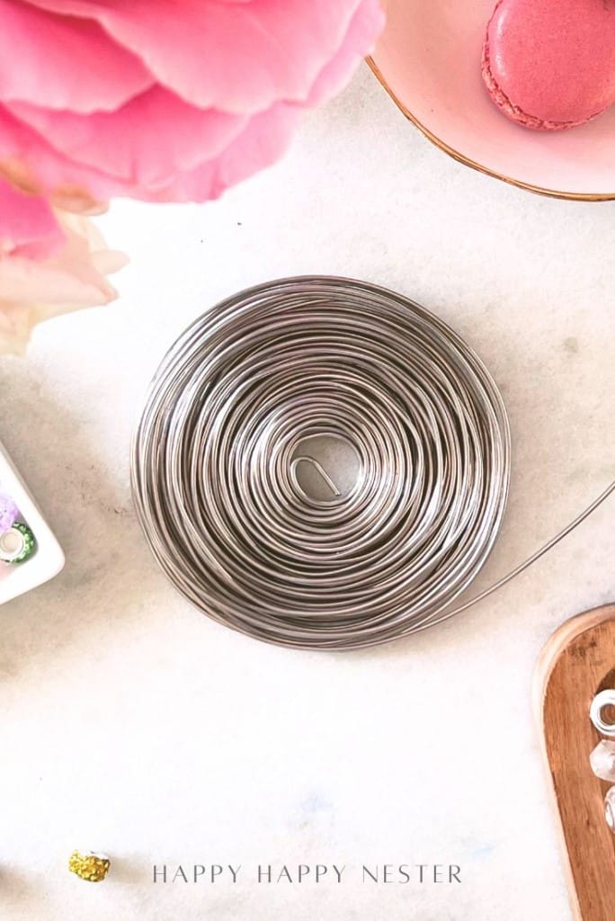 A coil of silver wire on a white surface, surrounded by a pink rose, a pink macaron on a plate, and decorative items; text at the bottom reads "HAPPY HAPPY NESTER.