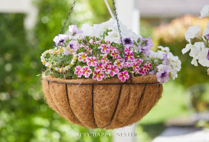 A hanging basket filled with blooming white, lavender, and pink flowers, set outdoors with a blurred green background. Accented with beaded garden stakes DIY touches, the basket is lined with coconut fiber.
