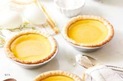 Four small lemon tarts in pie crusts sit alongside the elements of a Chinese egg custard tart recipe—eggs, flour, and a spoon—on a table with a napkin. The scene exudes a bright, inviting kitchen vibe.