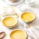 Four small lemon tarts in pie crusts sit alongside the elements of a Chinese egg custard tart recipe&mdash;eggs, flour, and a spoon&mdash;on a table with a napkin. The scene exudes a bright, inviting kitchen vibe.