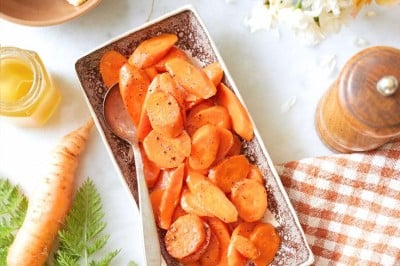 A rectangular dish with sliced cooked carrots on a marble surface, accompanied by a spoon. Nearby are a bowl of butter, honey, a fresh carrot with greens, a pepper grinder, a checkered napkin, and a bouquet of white flowers.