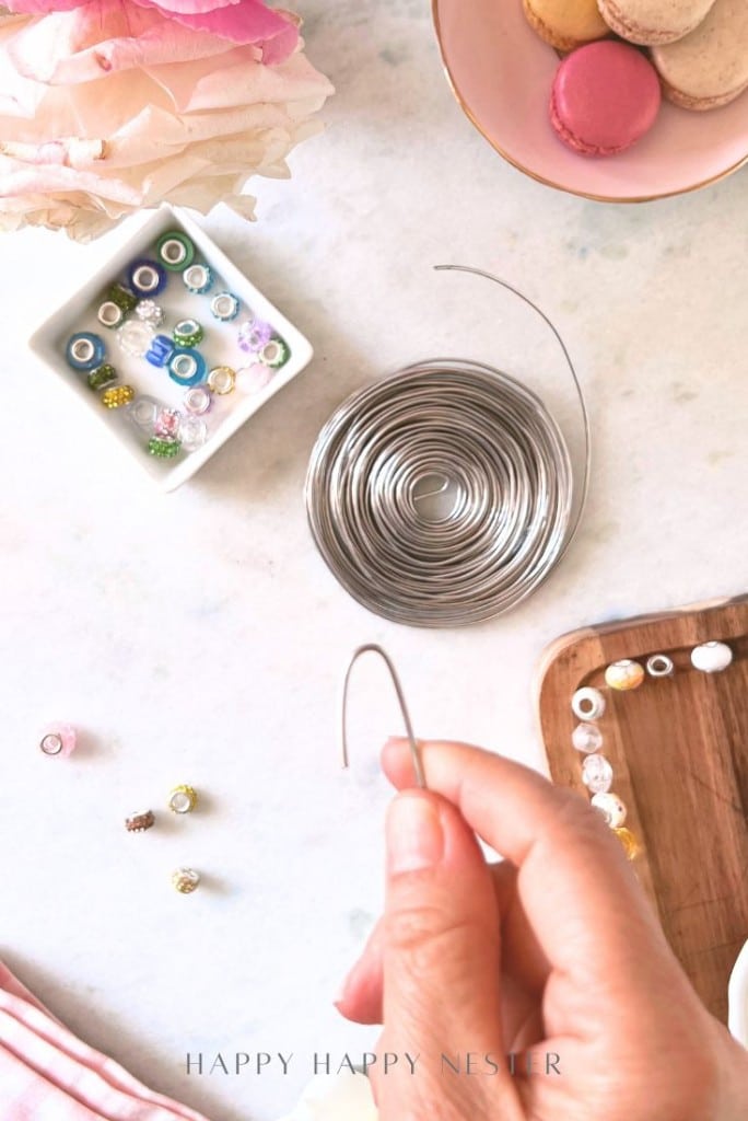 A hand holds a bendable wire above a table with a coiled wire, assorted beads in a dish and scattered nearby, a wooden tray, macarons on a plate, and pink flowers in the corner.