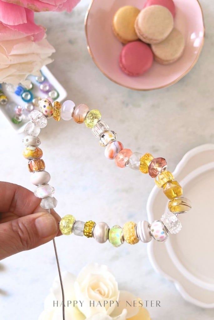 A hand holds a wire strung with colorful, assorted beads—perfect for beaded garden stakes DIY—above a table set with macarons, flowers, and jewelry-making supplies.