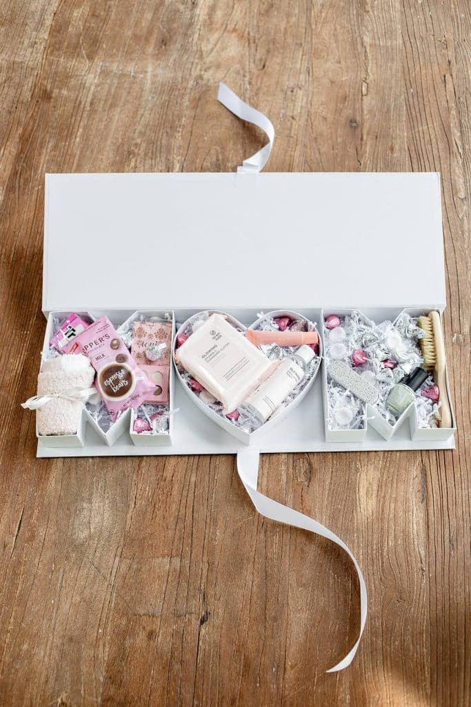 A white gift box with ribbon is open on a wooden table, revealing self-care items like lotions, face masks, soaps, and even beaded garden stakes for a touch of DIY garden decor—all neatly arranged in compartments.