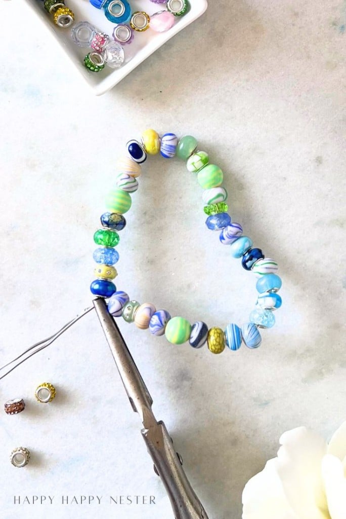A beaded bracelet with blue, green, and yellow beads is shaped into a heart with pliers on a white surface, perfect inspiration for beaded garden stakes DIY. Nearby are small beads in a tray and a white flower in the corner.