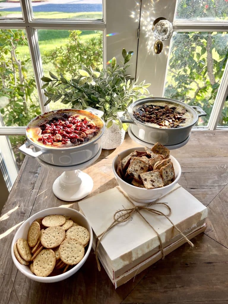 A wooden table by a sunny window displays two of my favorite side dishes—baked dips—alongside a bowl of crackers, crostini, a wrapped gift box, and a potted green plant. Sunlight streams through the glass, casting bright reflections.