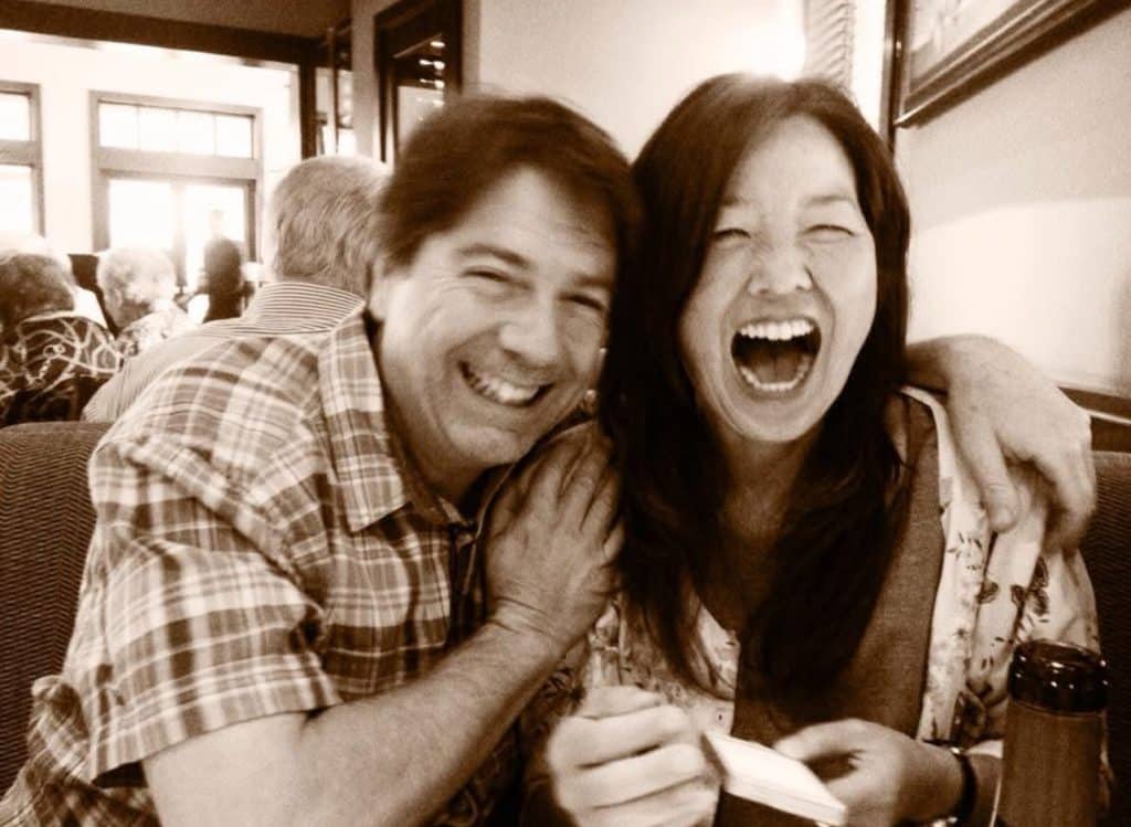 A man and woman sit close together in a restaurant booth, both laughing joyfully. The man has his arm around her shoulders; she is holding a cup, perhaps enjoying one of my favorite side dishes. The sepia tone adds a warm, nostalgic feel.