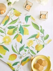 A flat lay of a lemon-patterned napkin, yellow plate with cookies, two lit candles in golden holders, and white flowers on a white tablecloth.