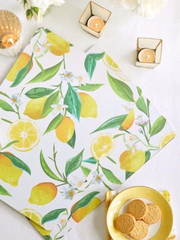 A flat lay of a lemon-patterned napkin, yellow plate with cookies, two lit candles in golden holders, and white flowers on a white tablecloth.
