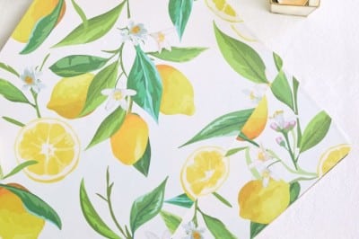A flat lay of a lemon-patterned napkin, yellow plate with cookies, two lit candles in golden holders, and white flowers on a white tablecloth.
