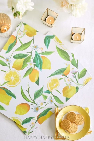 A flat lay of a lemon-patterned napkin, yellow plate with cookies, two lit candles in golden holders, and white flowers on a white tablecloth.