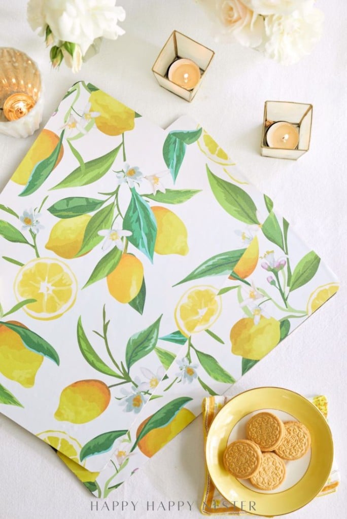 A flat lay of a lemon-patterned napkin, yellow plate with cookies, two lit candles in golden holders, and white flowers on a white tablecloth.