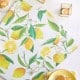 A flat lay of a lemon-patterned napkin, yellow plate with cookies, two lit candles in golden holders, and white flowers on a white tablecloth.