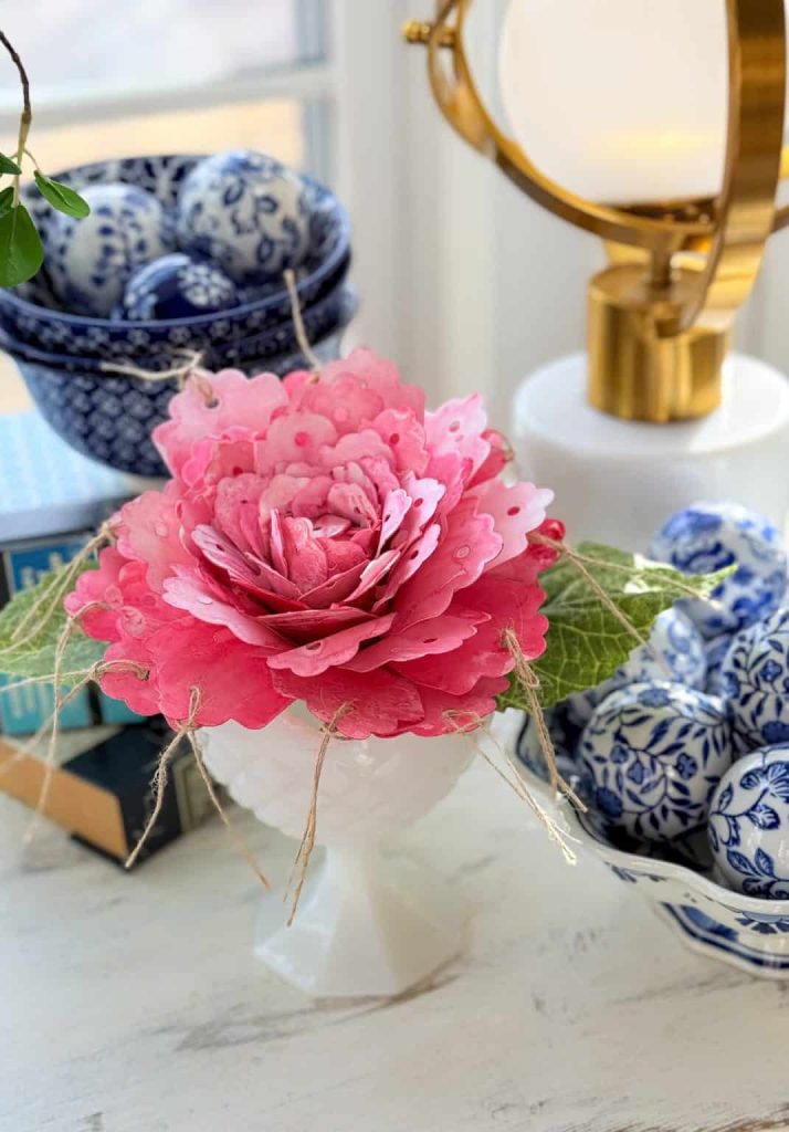 A pink paper flower, one of my favorite flower crafts, with green leaves sits in a white vase, surrounded by blue-and-white patterned bowls, decorative eggs, and books on a light-colored table near a gold lamp.