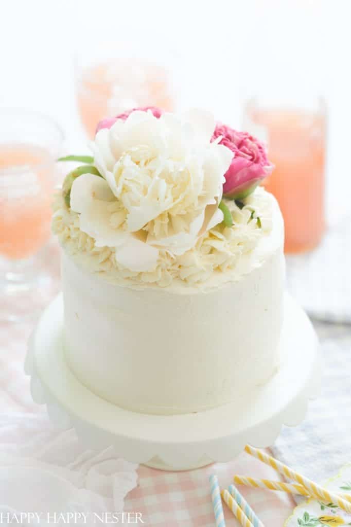 A white frosted cake decorated with large white and pink flowers, inspired by my favorite flower inspiration, sits on a white cake stand. In the background are glasses of pink beverage and pastel-striped straws on a checkered tablecloth.