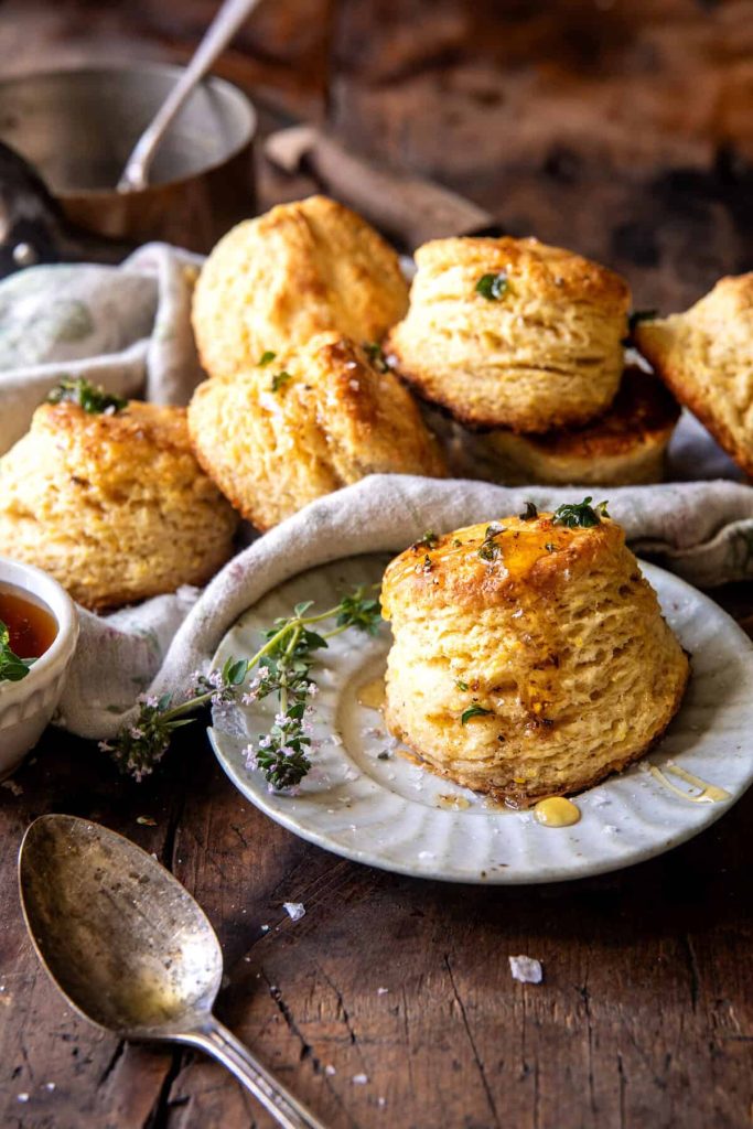 Fluffy golden biscuits are stacked on a rustic plate and wooden table, with honey drizzled on top and fresh herbs as garnish—one of my favorite side dishes. A vintage spoon and a small bowl of honey are nearby.
