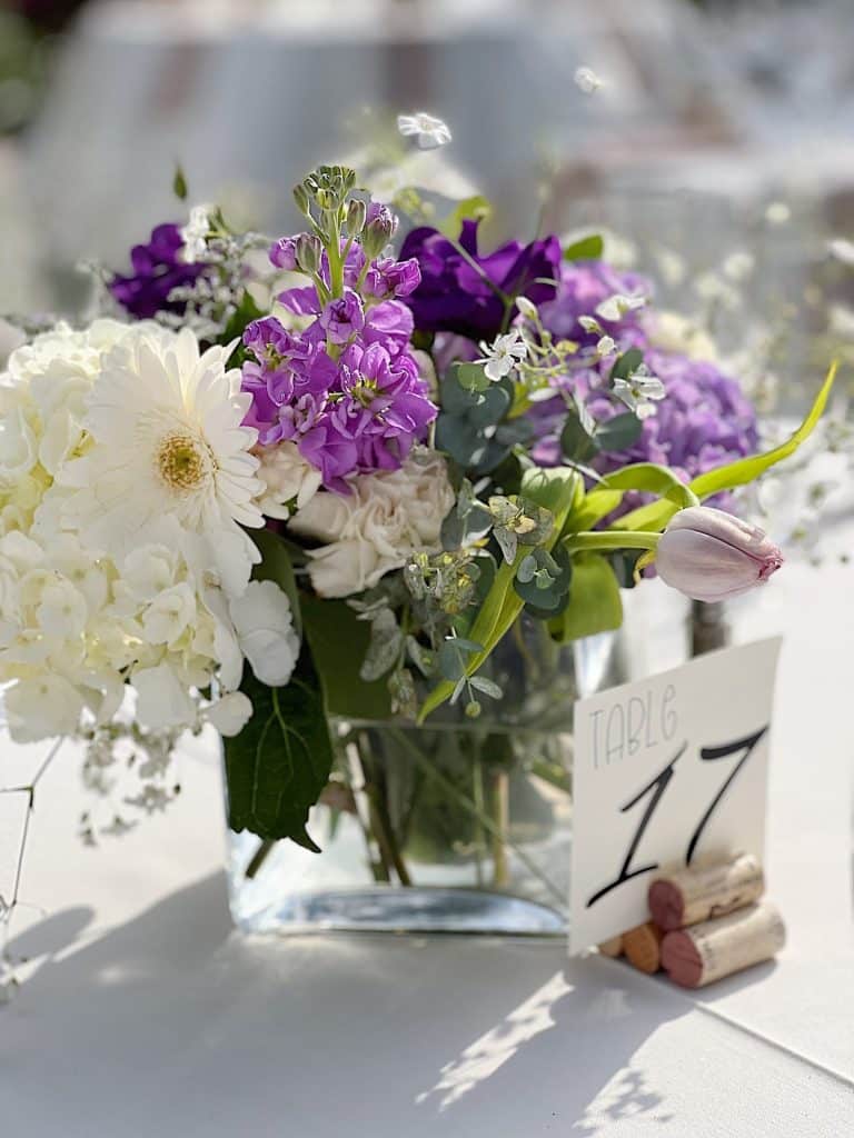 A clear glass vase filled with white and purple flowers—my favorite flower inspiration—sits on a table. Next to it is a card reading "Table 17," propped up by several wine corks, with a softly blurred background.