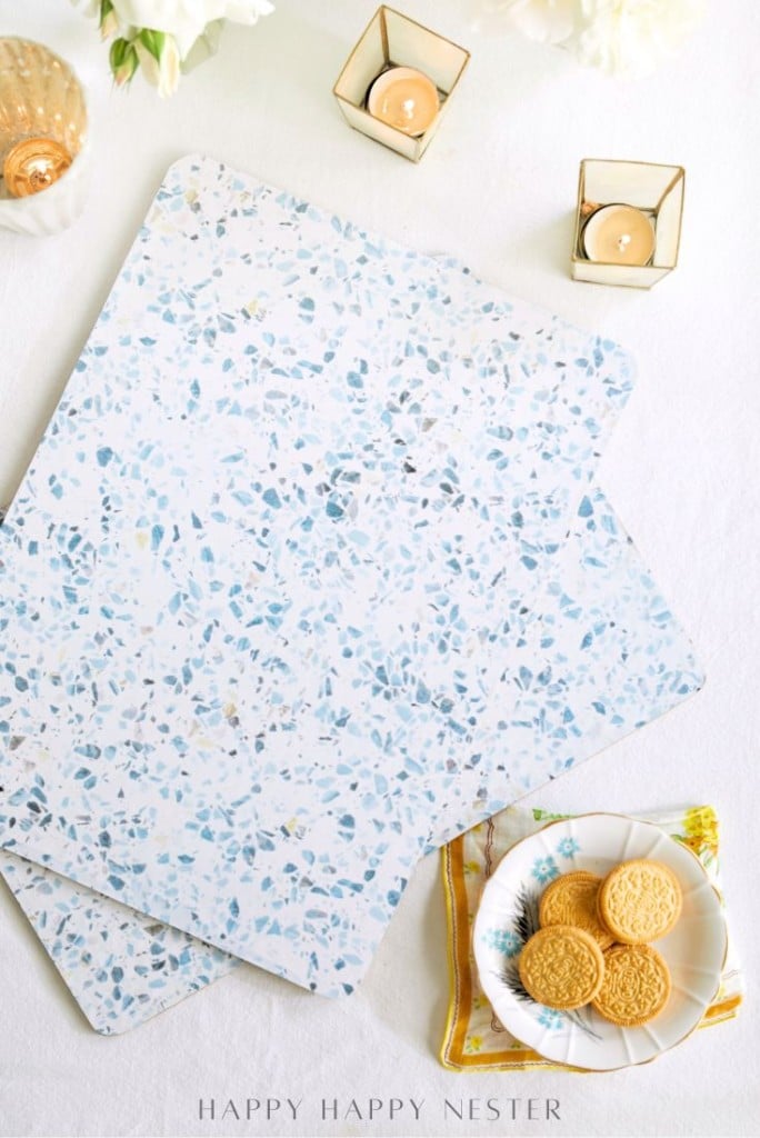 Two terrazzo-patterned, easy corkboard placemats in blue and white tones sit on a table. Nearby are a plate with cookies, a yellow napkin, and two lit candles in small glass holders. Flowers are partially visible at the top.