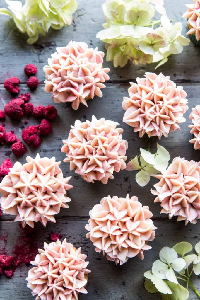 Several cupcakes with pale pink, flower-like frosting are arranged on a rustic wooden surface, surrounded by dried raspberries and green hydrangea flowers—my favorite flower inspiration for a charming dessert display.