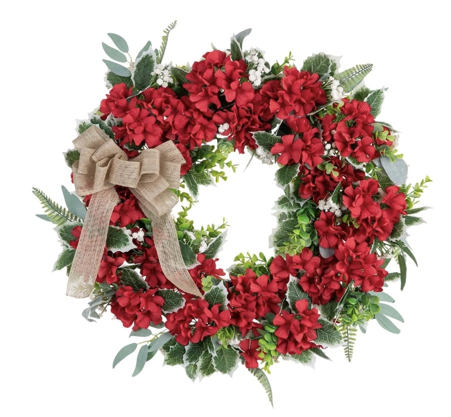 A festive 4th of July wreath with vibrant red flowers, green leaves, and sprigs of white, adorned with a large burlap bow on the left side, set against a white background.
