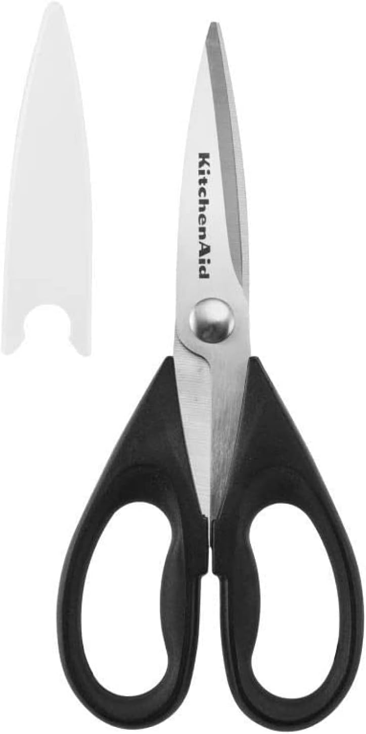 A pair of KitchenAid kitchen scissors with black handles and stainless steel blades, shown alongside a white blade cover—an essential tool when learning how to make a tender juicy steak at home.