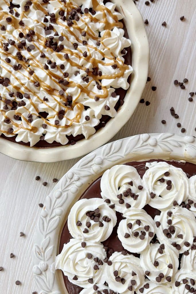 Two chocolate pies topped with whipped cream, chocolate chips, and caramel drizzle on a light wooden surface, perfect for trying as a dessert recipe. Extra chocolate chips are scattered around for a delicious finishing touch.