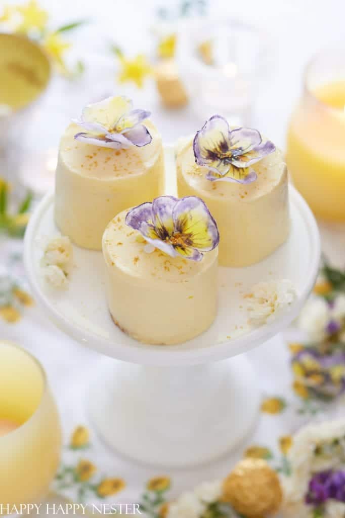 Three small, round lemon cakes topped with purple and yellow edible flowers—my favorite flower inspiration—are displayed on a white cake stand, surrounded by candles, drinks, and scattered flower petals.