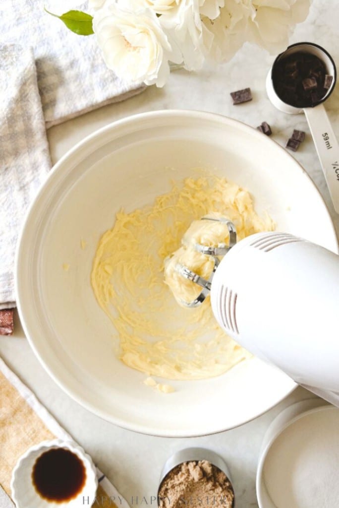 A hand mixer blends butter in a white mixing bowl on a marble countertop, surrounded by sugar, chocolate chunks, and vanilla—perfect ingredients for your favorite chocolate chip bars recipe—plus a towel and white flowers in a vase.